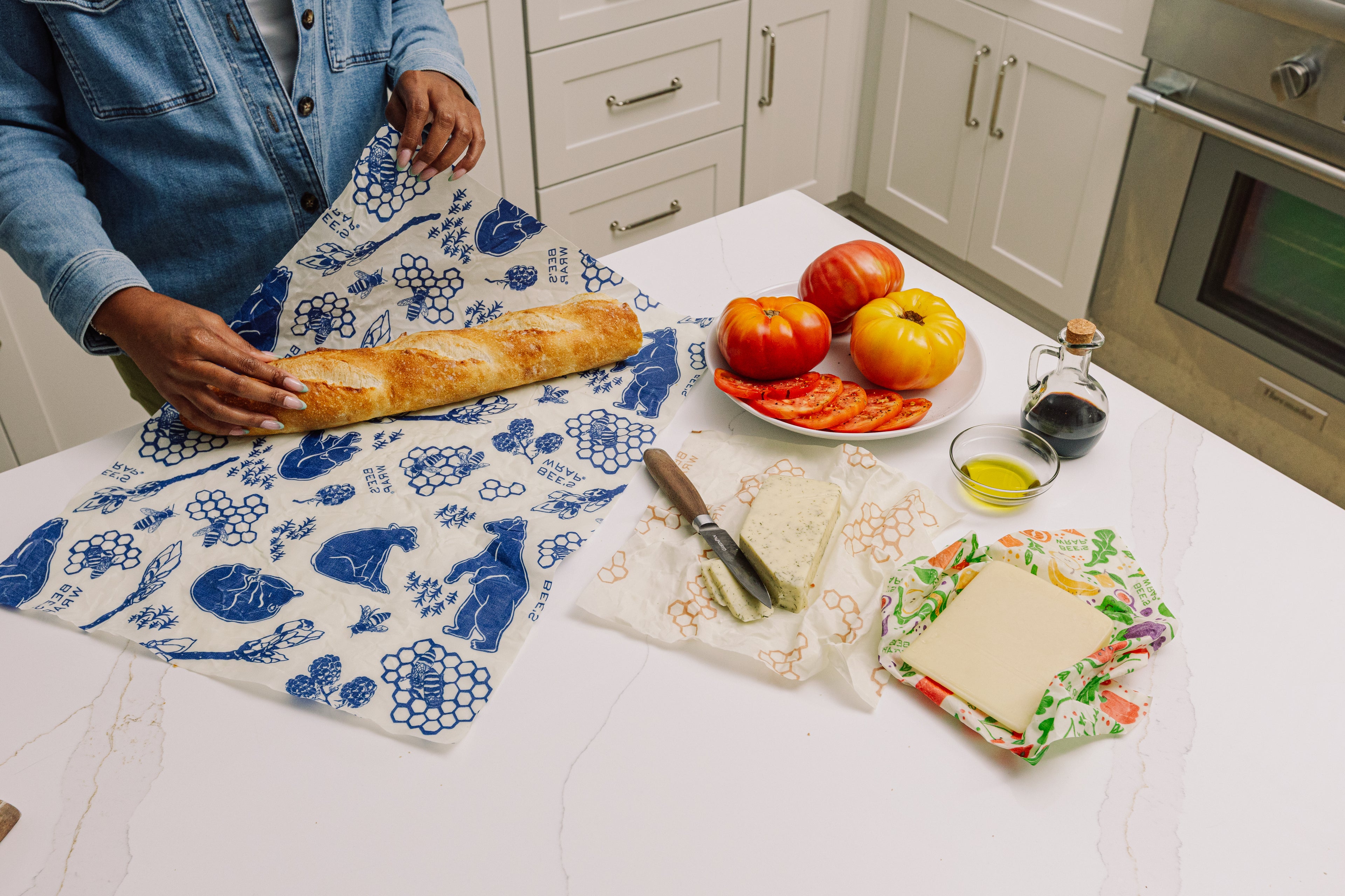 Person wrapping a baguette in Bees and Bears Roll on a kitchen counter with bread, tomatoes, and other items